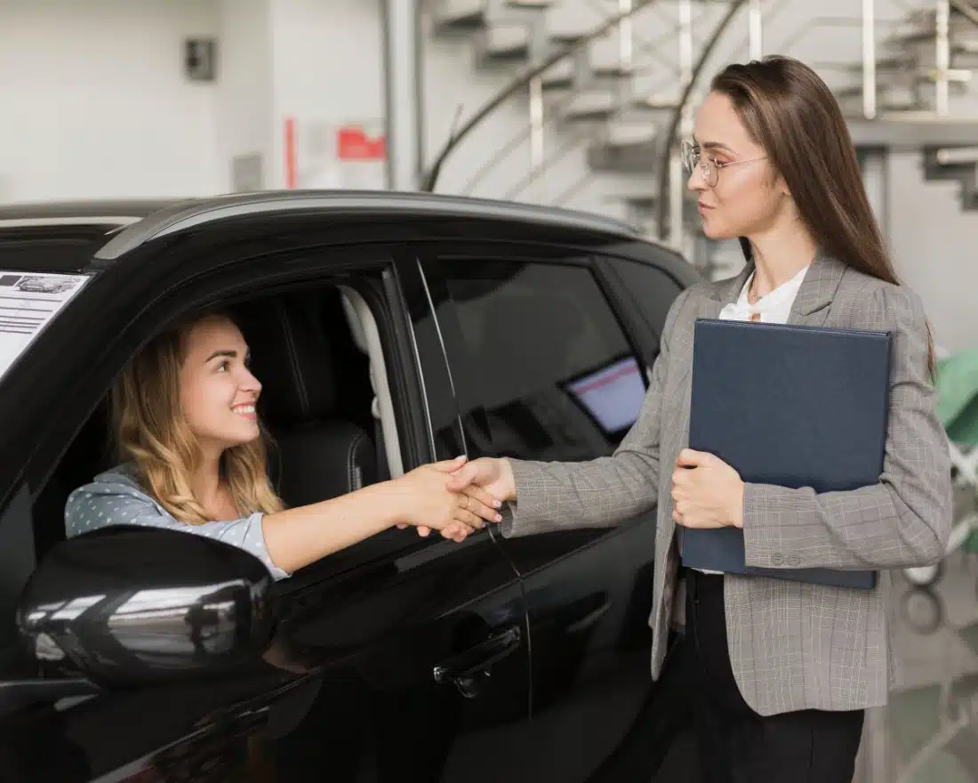 A smiling woman inside a black car shakes hands with a professional in a gray blazer, symbolizing the easy and friendly title pawn process in Jasper, GA.