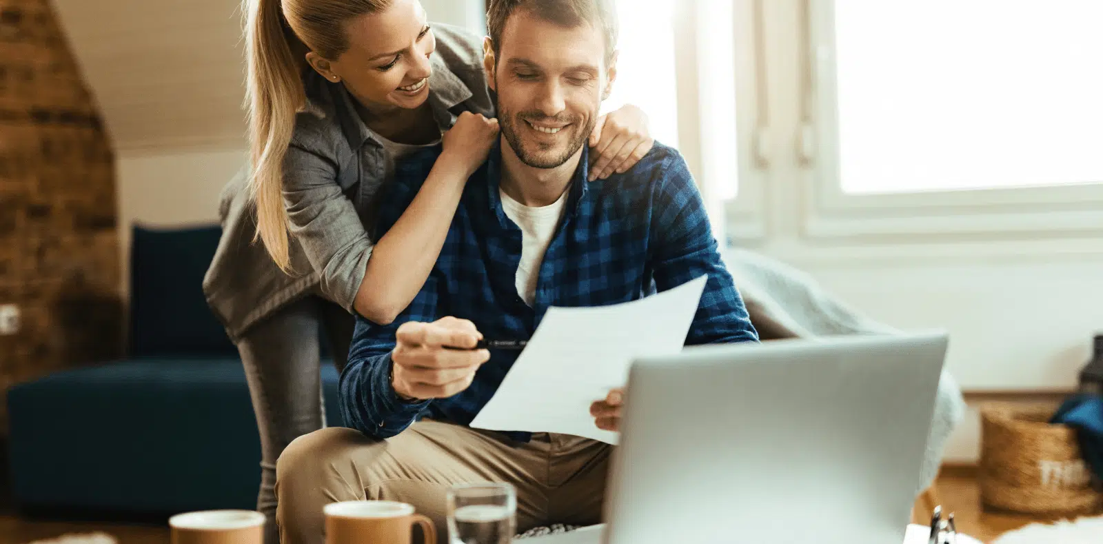 Two hands hold open a black leather wallet stuffed with $100 bills, with more cash spread loosely on a surface nearby