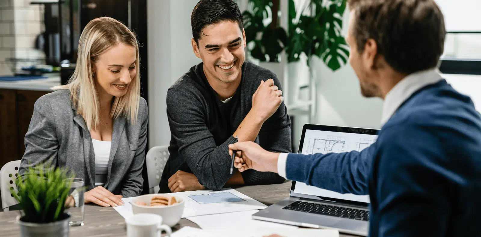 A bearded man with glasses signs paperwork at a table while a woman beside him and a smiling blonde advisor in a blazer look on